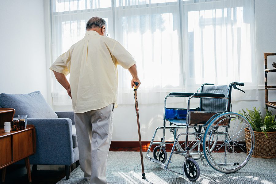 elderly man walks with a cane across the room to a wheelchair - transitioning home after a hospital stay concept