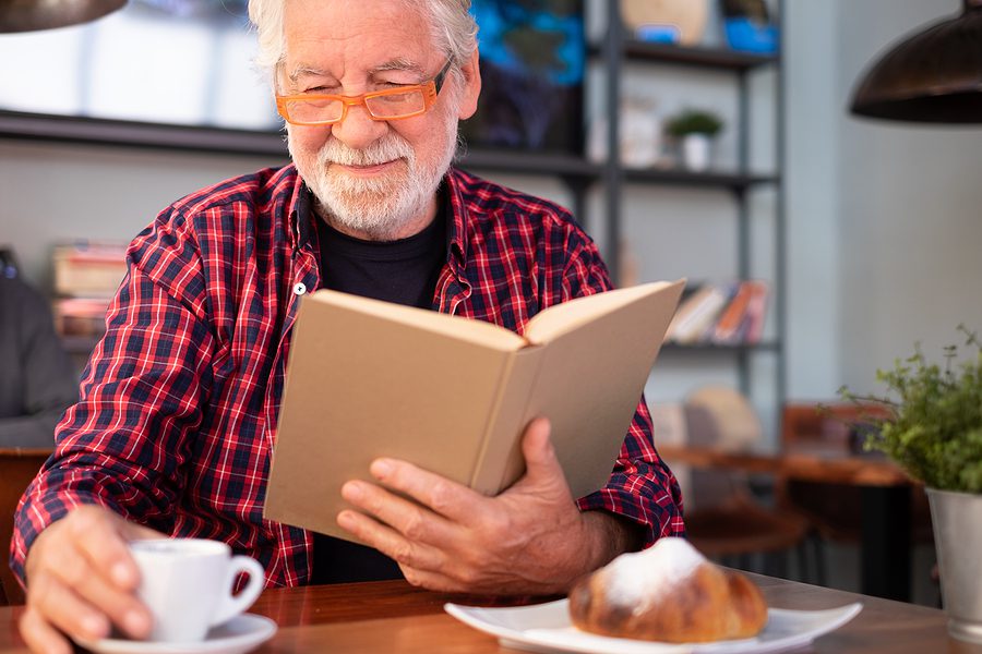 bearded man with white hair drinking coffee and reading a book during his morning meal