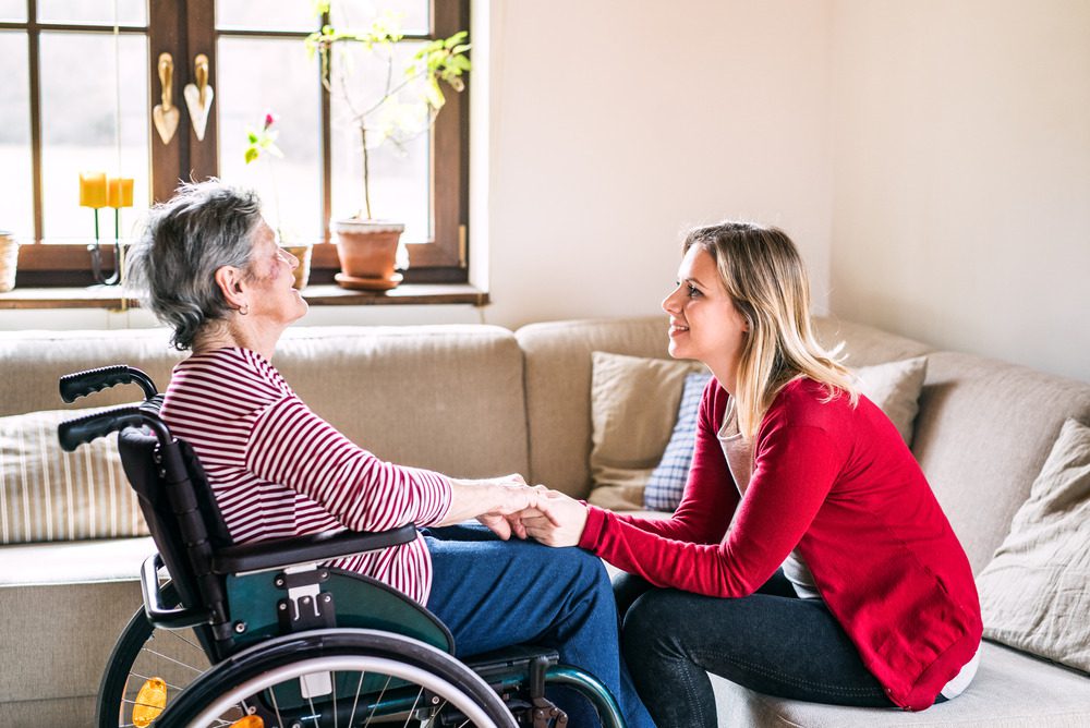 older woman in wheelchair speaking with younger caretaker on the couch