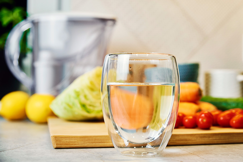 glass of water in front of filter pitcher and a counter full of vegetables - hydration and incontinence concept