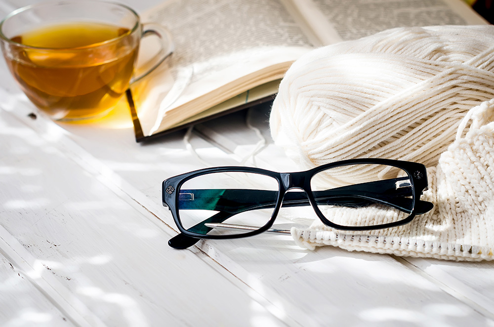 reading glasses on table with a book, a cup of tea, and knitting yarn
