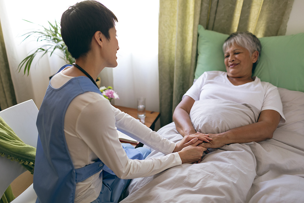 bedridden older woman resting in bed while holding her aide's hand